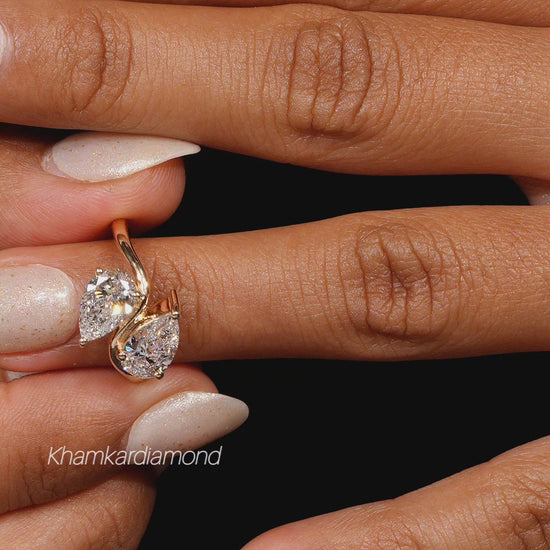 Engagement ring with yellow metal bypass band, two pear-shaped clear gemstones, worn on finger, close-up front view
