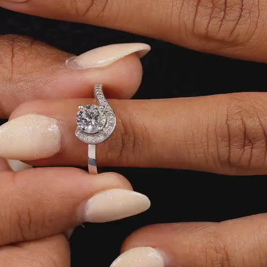 Engagement ring with round center stone, white metal swirl halo design, pavé accent band, worn on finger, front view
