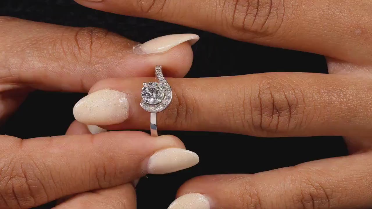 Engagement ring with round center stone, white metal swirl halo design, pavé accent band, worn on finger, front view
