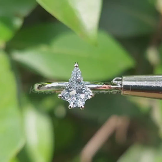 Engagement ring video with white metal band, kite-shaped clear center stone in prong setting, front view against green leaves
