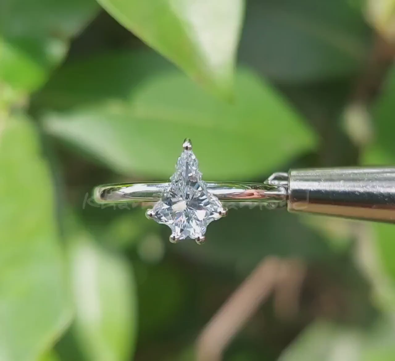 Engagement ring video with white metal band, kite-shaped clear center stone in prong setting, front view against green leaves
